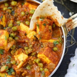 A close up shot of Matar Paneer in a copper pan with a piece of roti scooping the paneer.