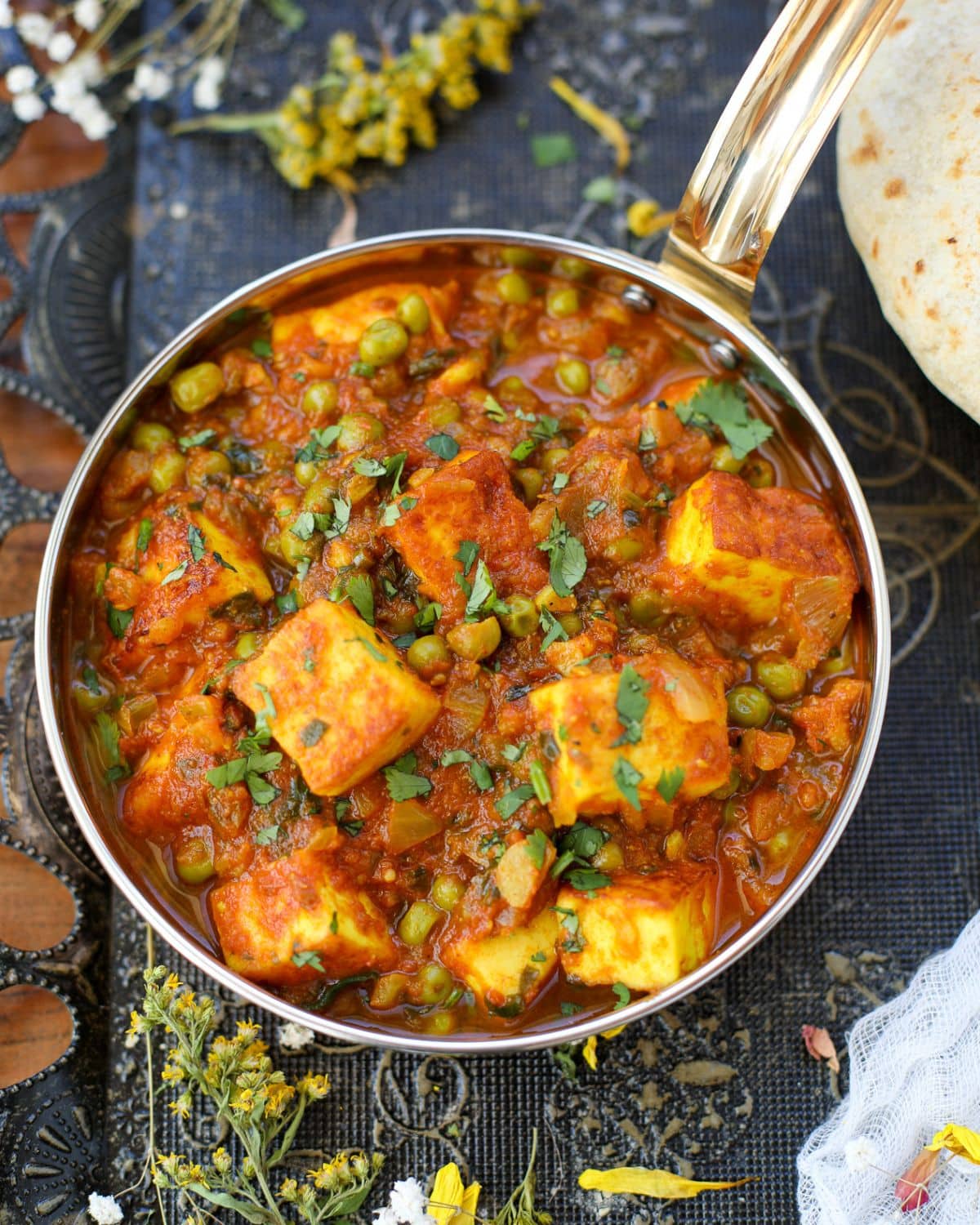 A small copper pan containing matar paneer surrounded by yellow and white flowers with roti on the upper right corner.