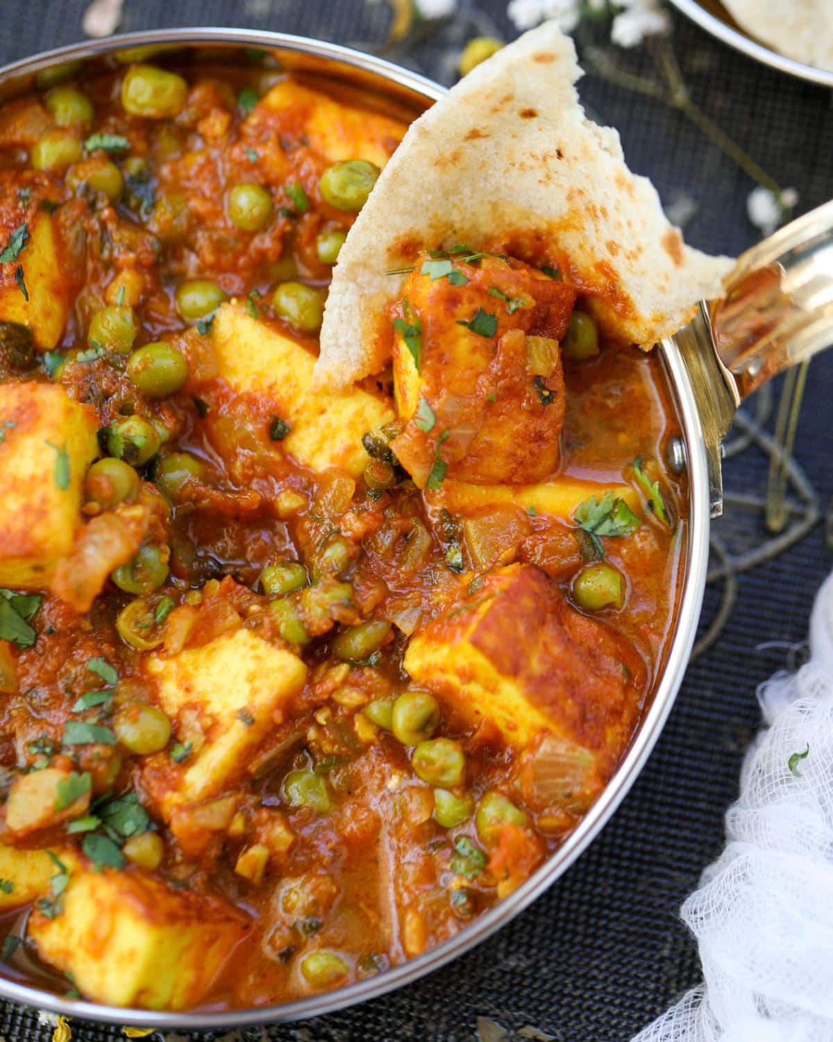 A close up shot of Matar Paneer in a copper pan with a piece of roti scooping the paneer.