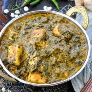 A serving bowl of creamy palak paneer with green chilies and white flowers in the background.