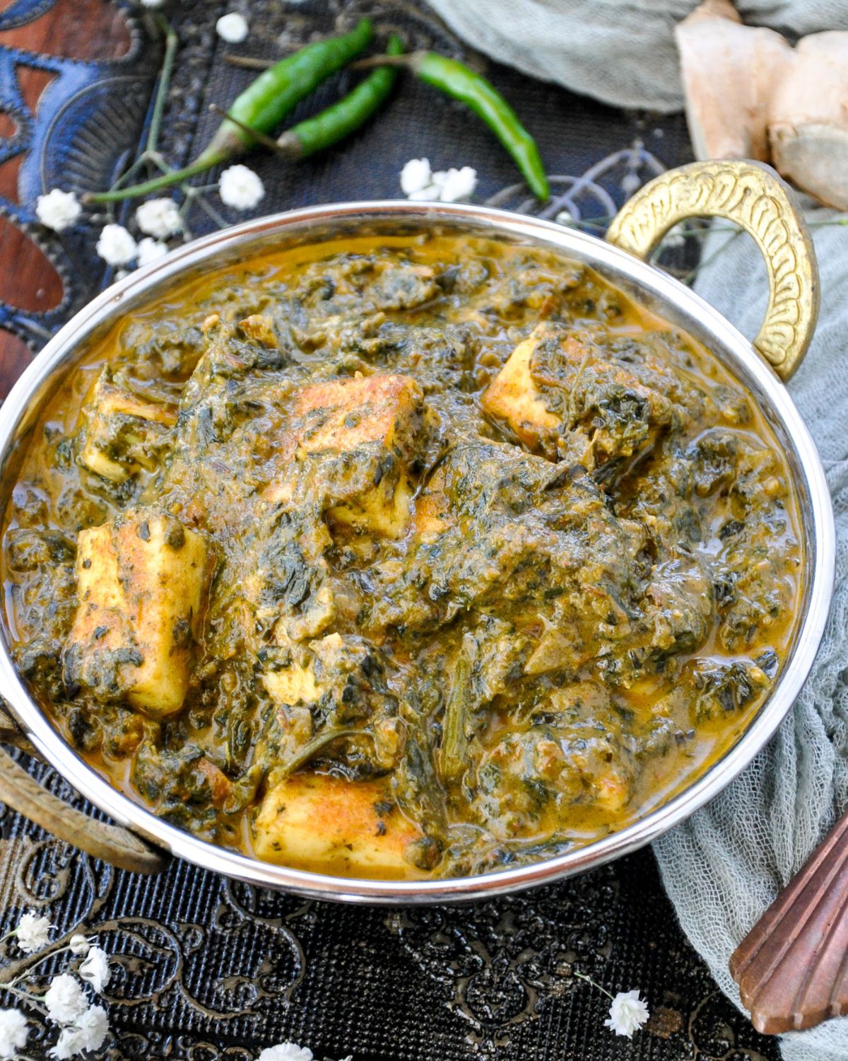 A serving bowl of creamy palak paneer with green chilies and white flowers in the background.