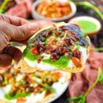 Piece of Masala Papad topped with yogurt, veggies, chutneys, and spices with veggies and green chutney in the background.