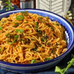 A bowl of chili garlic noodles garnished with scallions with a jar of chili crunch and a small plant in the back.