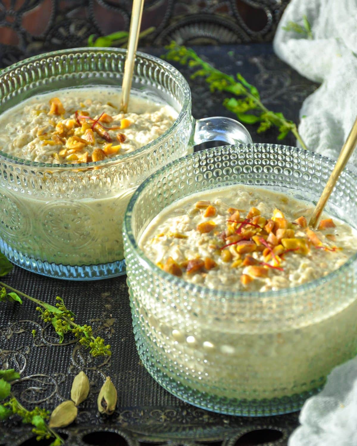 Pistachio Overnight Oats in a fancy glass mug with 2 serving gold spoons topped with chopped pistachios and a few saffron threads.
