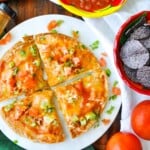 Cheese and vegetable pizza on a round cutting board, reminiscent of a Taco Bell Mexican Pizza, topped with diced tomatoes and green onions. Nearby are bowls of salsa and blue corn tortilla chips, with fresh tomatoes on the side.