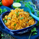 Bowl of Mexican Rice with a large blue serving spoon and a tomato and lime in the background.