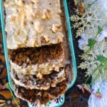 A loaf of date walnut coffee cake topped with coffee buttercream and garnished with walnuts, there is a cup of coffee on the upper right and a couple snowflake ornaments towards the right.