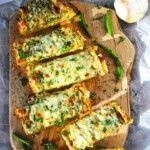 Spicy cheesy garlic bread sliced intoa pieces and placed on a wooden cutting board surrounded by a few green chilies and garnished with cilantro.