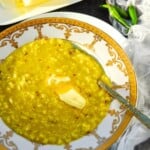 A bowl of plain khichdi flavored with cumin seeds topped with butter slowly melting, and to the side are three Thai green chilies.