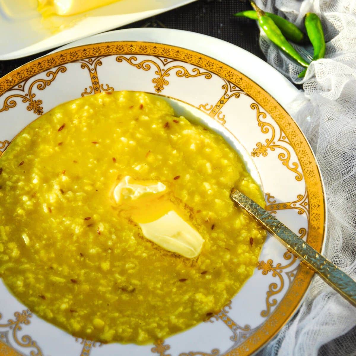 A bowl of plain khichdi flavored with cumin seeds topped with butter slowly melting, and to the side are three Thai green chilies.