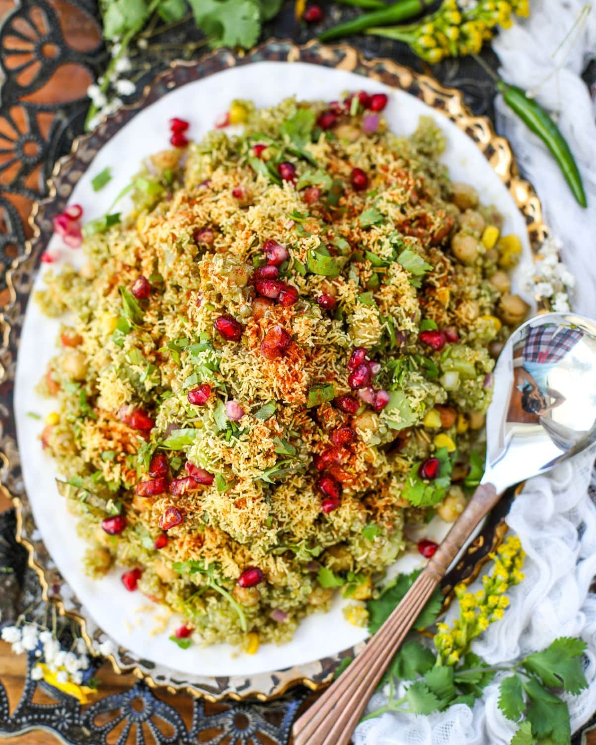An overhead shot of Quinoa Bhel Salad on a white plate with gold trim with a copper spoon surrounded by yellow and white flowers. The dish is garnished with cilantro, sev, and pomegrante.
