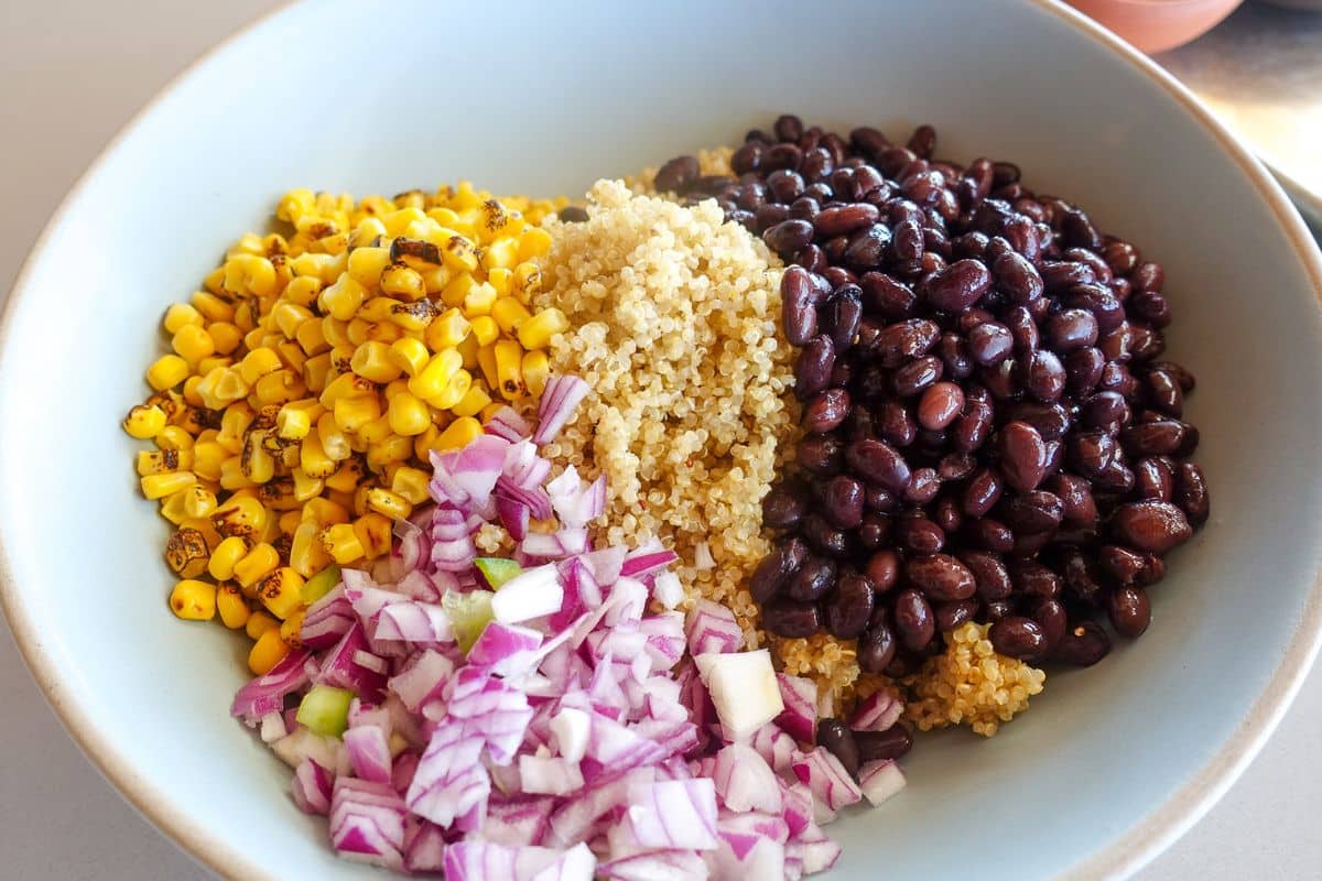 A bowl containing grilled corn, quinoa, black beans, and chopped red onions arranged in separate sections.