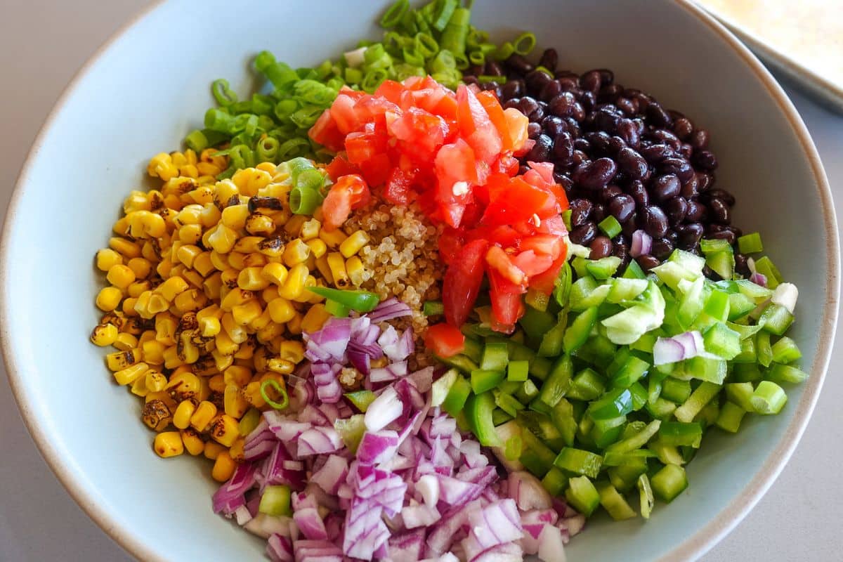 A bowl with sections of black beans, diced tomatoes, grilled corn, chopped green onion, diced green bell pepper, and chopped red onion.