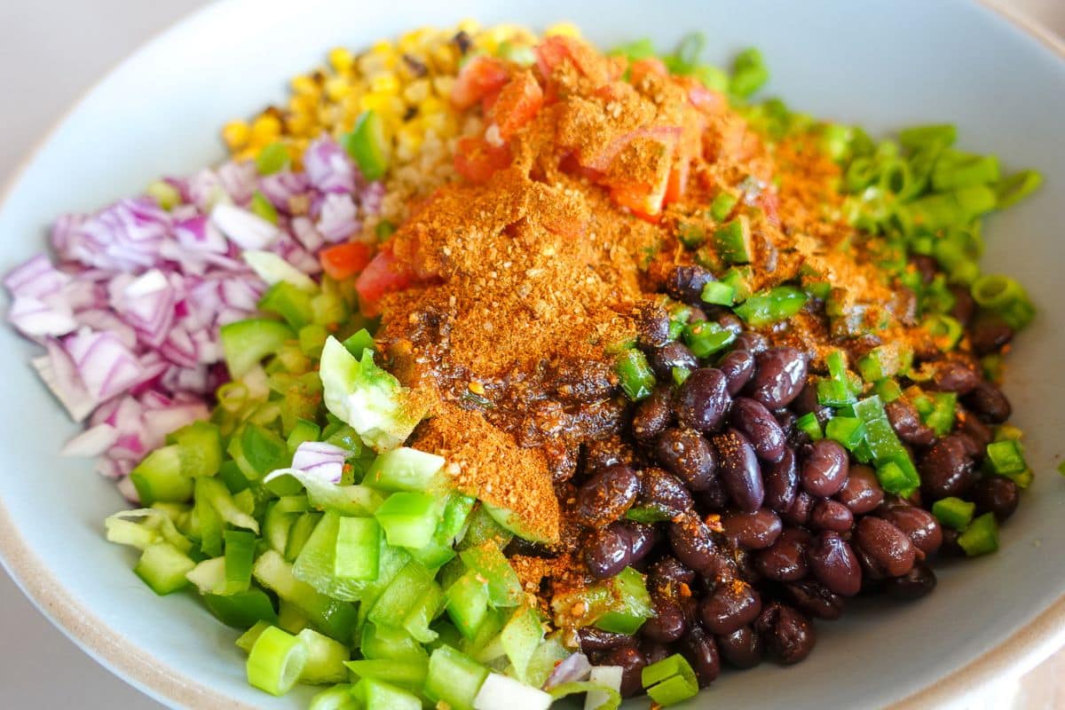 A bowl filled with chopped red onions, green bell peppers, black beans, diced tomatoes, corn, green onions, and a generous amount of taco seasoning.