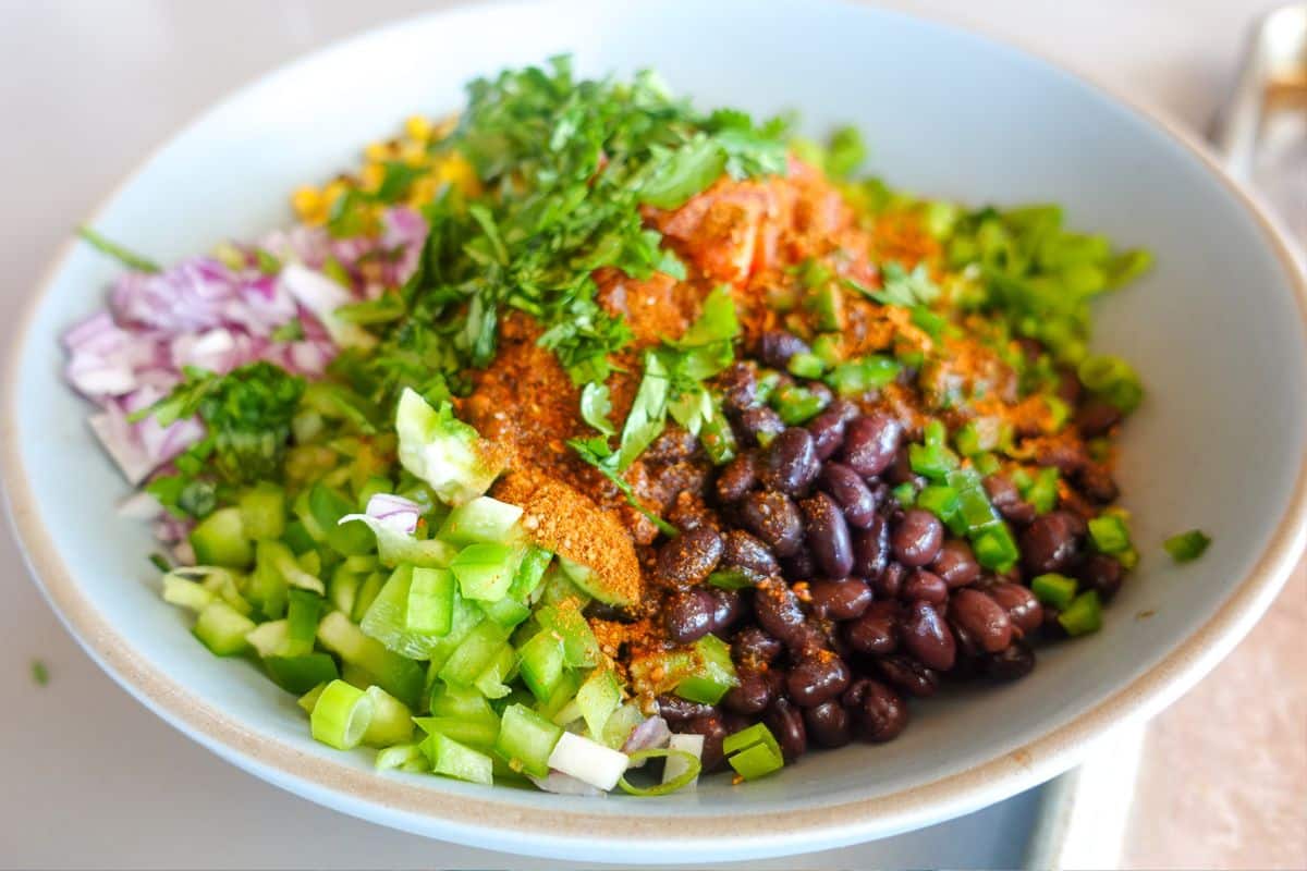 A bowl filled with black beans, chopped green bell pepper, red onion, corn, green onions, fresh cilantro, and various spices.