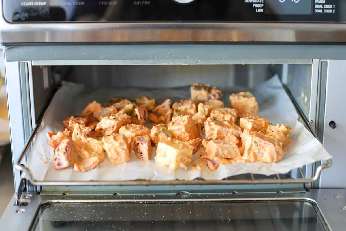 Cubes of paneer coated in a creamy marinade are arranged on parchment paper inside a countertop toaster oven.