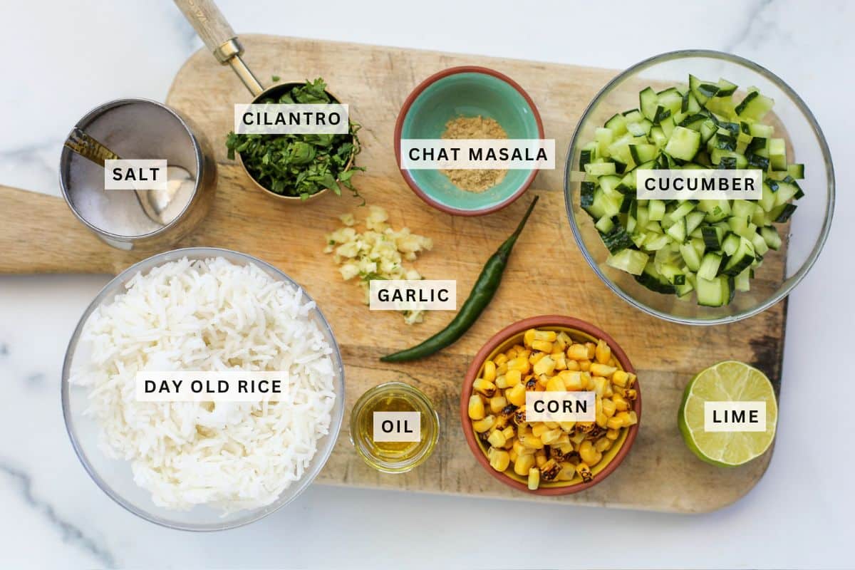 A wooden board displays bowls of day-old rice, corn, chopped cucumber, garlic, cilantro, chat masala, salt, oil, a green chili, and a halved lime.