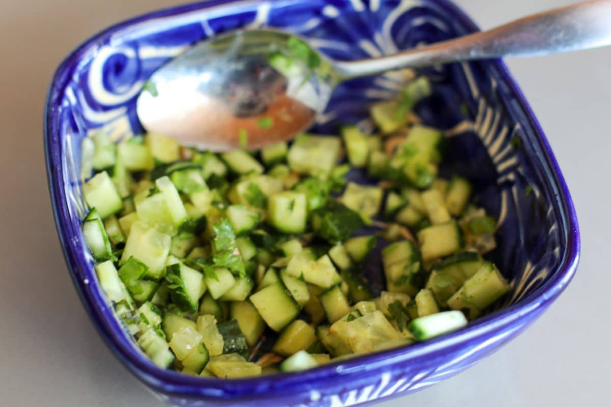 A blue bowl containing a spoon wtih chopped cucumber, chat masala, and lime juice.