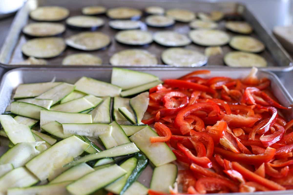 Sliced zucchini and red bell peppers arranged on a baking tray with sliced eggplant on a second tray in the background.