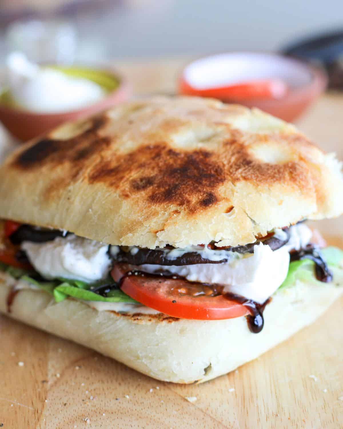 A grilled vegetable panini with burrata, tomato, lettuce, roasted veggies, and balsamic glaze on focaccia bread sits on a wooden surface.