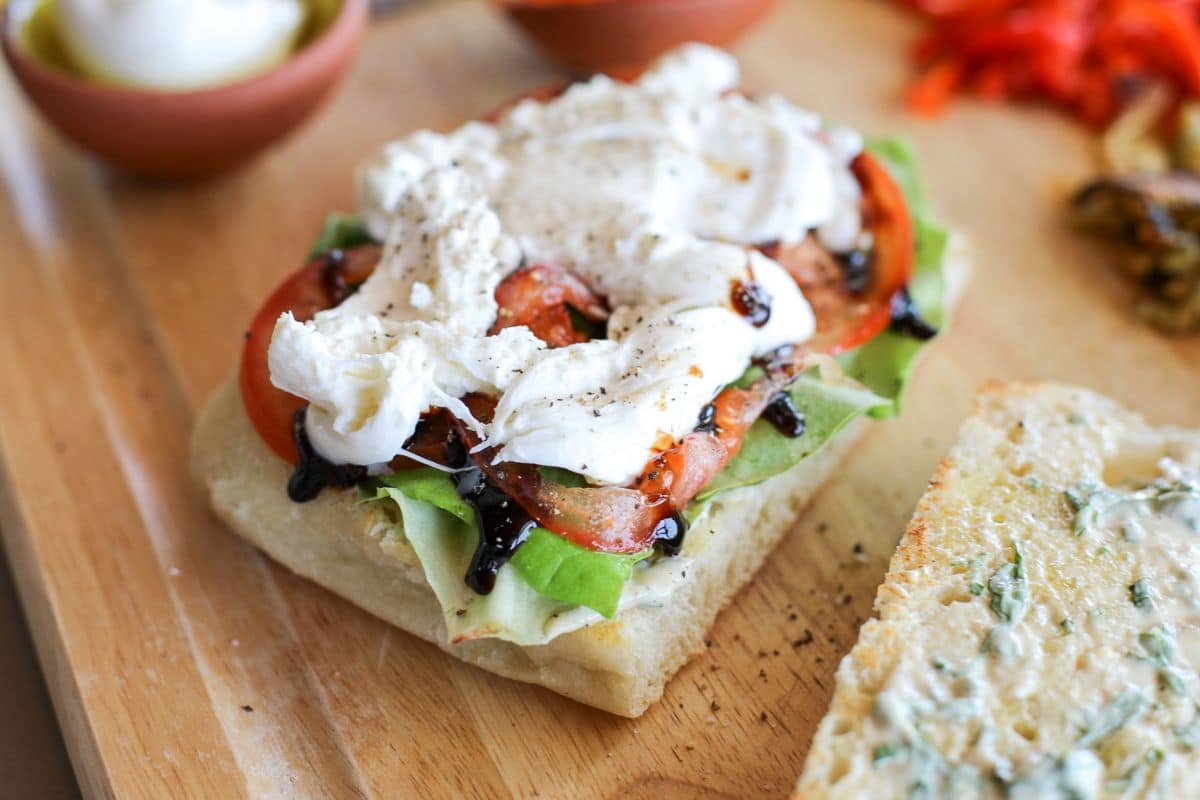 An open sandwich on ciabatta bread topped with lettuce, tomato slices, mozzarella cheese, and balsamic glaze, on a wooden board with other food items in the background.