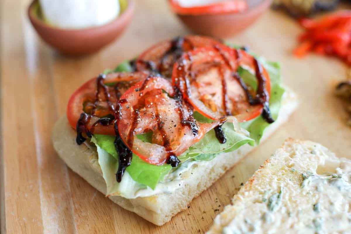 Open sandwich on a wooden surface with sliced tomatoes, lettuce, and balsamic glaze on ciabatta bread; other sandwich ingredients are visible in the background.