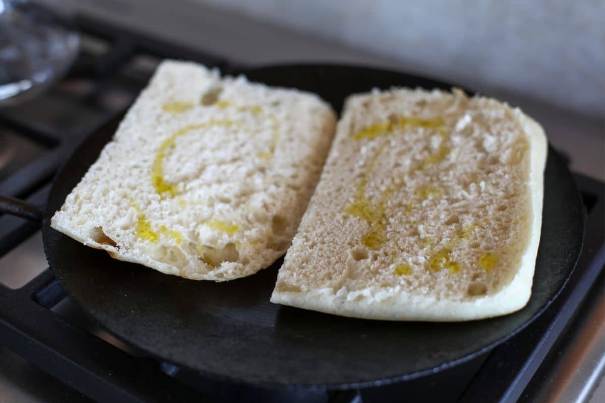 Two halves of a sandwich bun with a drizzle of olive oil are placed face up on a skillet, ready to be toasted.