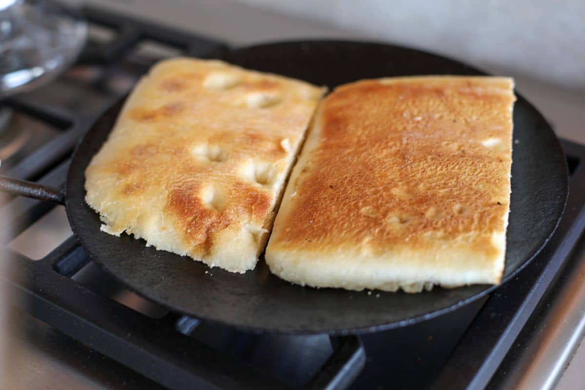 Two rectangular pieces of flatbread are being toasted on a round cast iron pan over a stove.