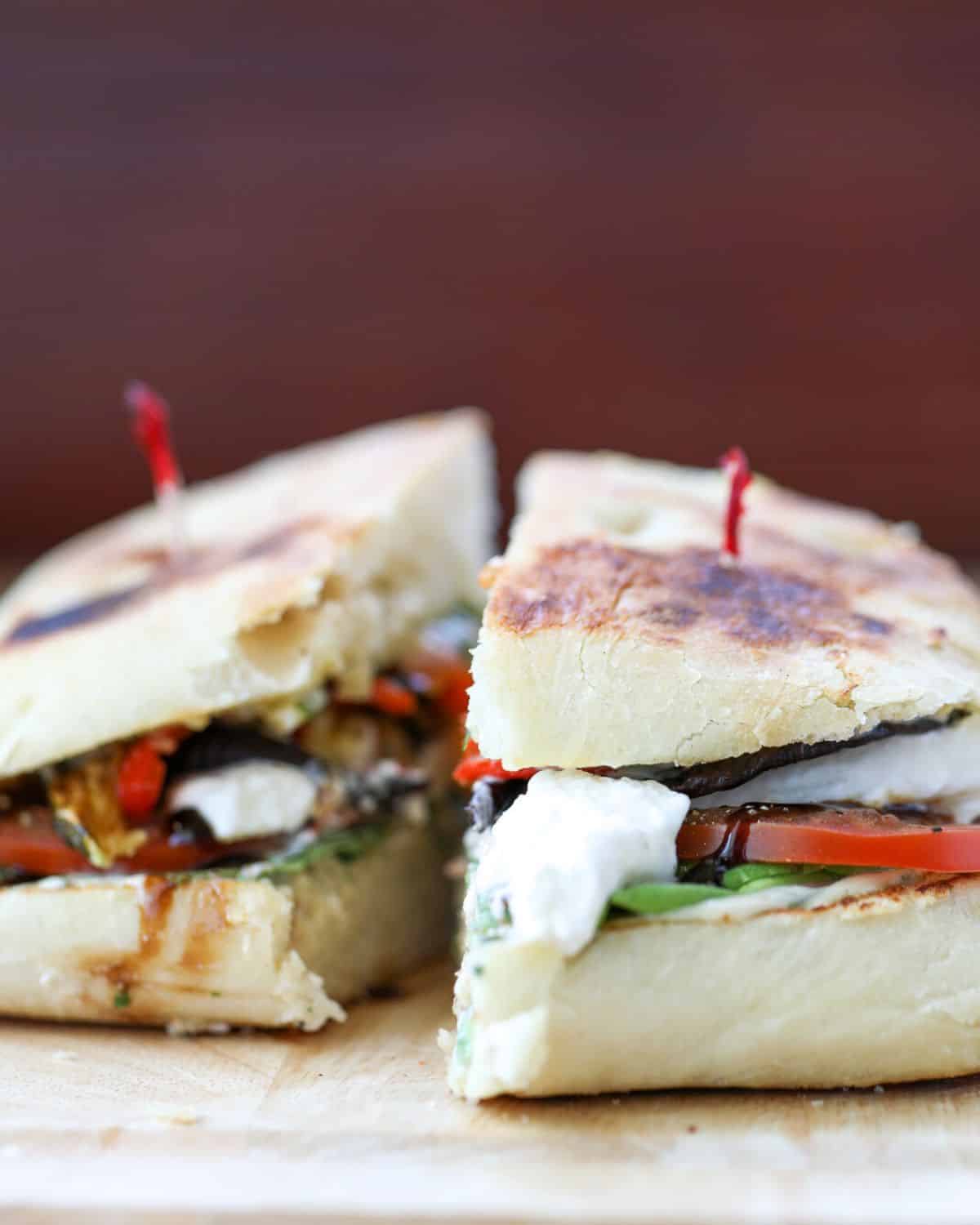 A grilled sandwich cut in half, filled with vegetables, cheese, and sauces, served on a wooden surface with red toothpicks securing each half.