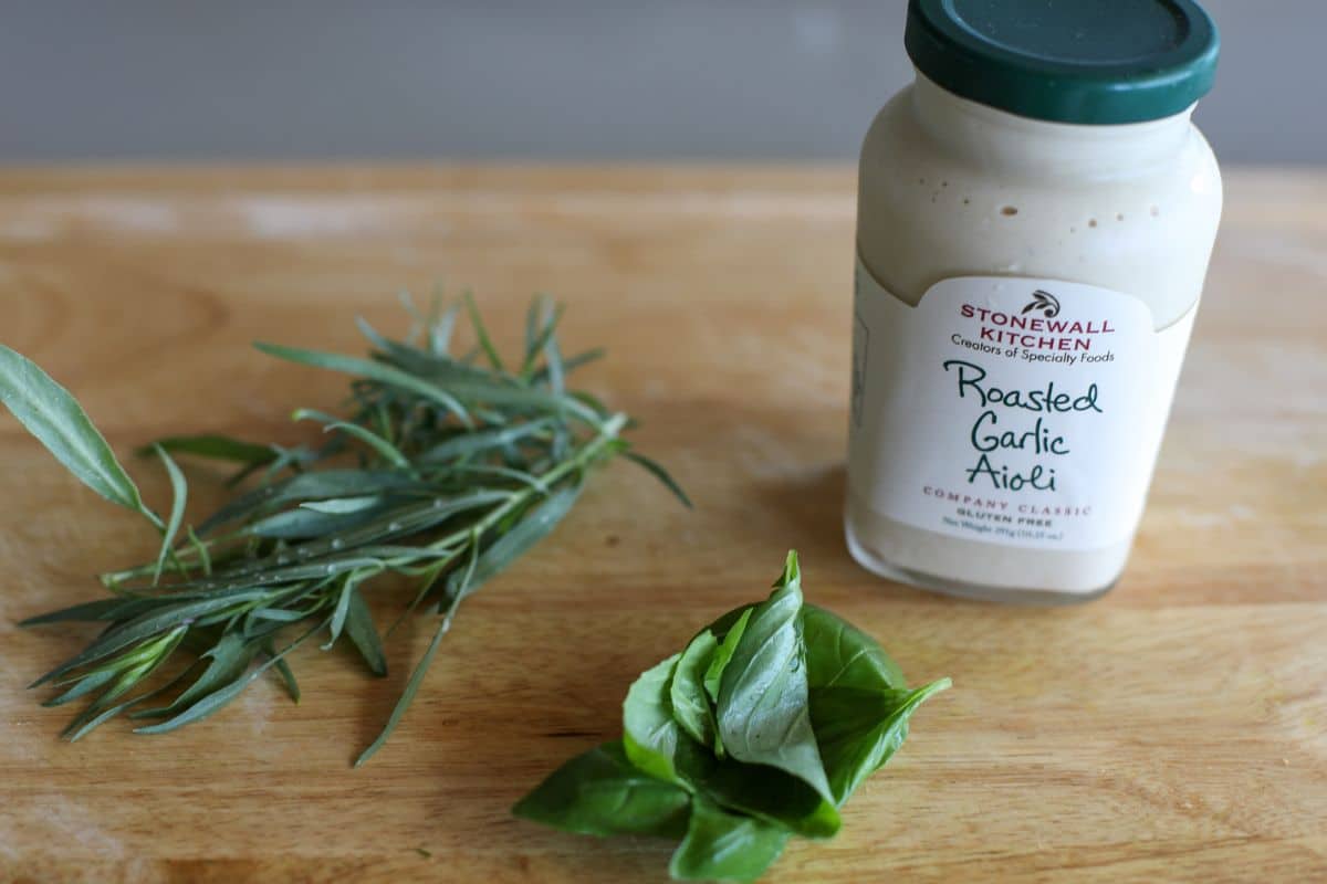A jar of Stonewall Kitchen Roasted Garlic Aioli sits on a wooden surface next to fresh basil leaves and a sprig of tarragon.