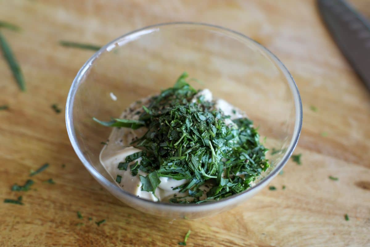 A glass bowl containing a mixture of aioli and chopped fresh herbs sits on a wooden surface.