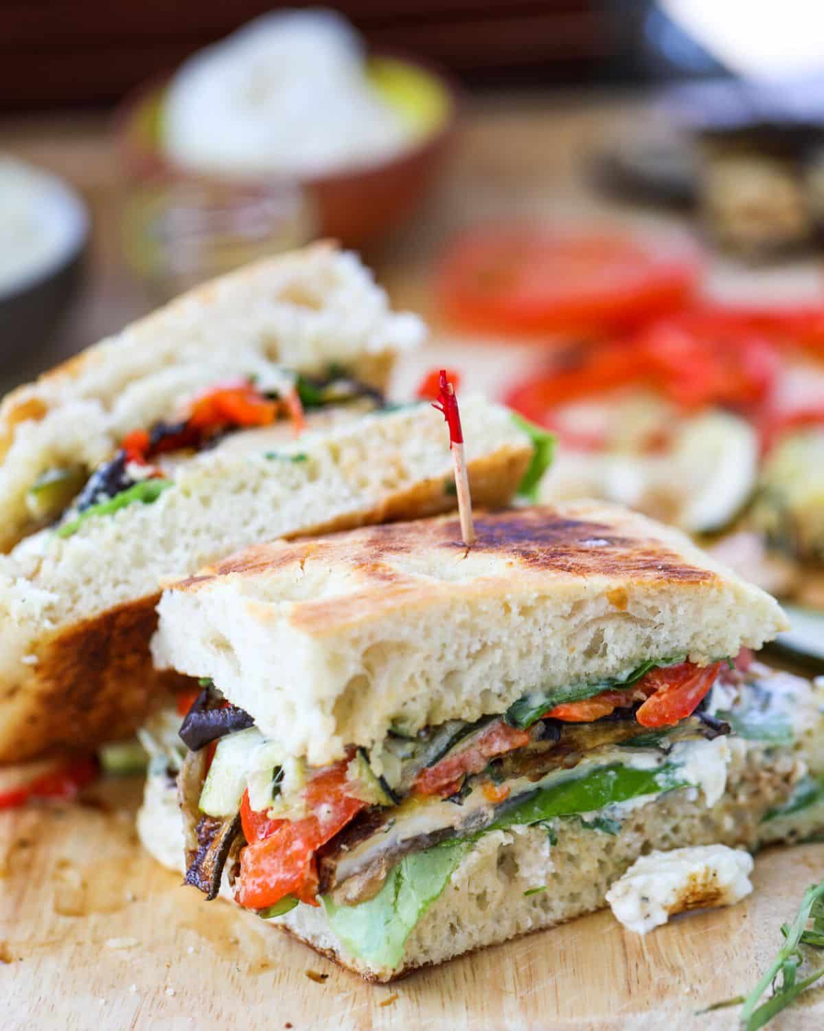 A close-up of a vegetable sandwich on focaccia bread, cut in half and stacked, showing layers of grilled vegetables and burrata with a toothpick holding it together.