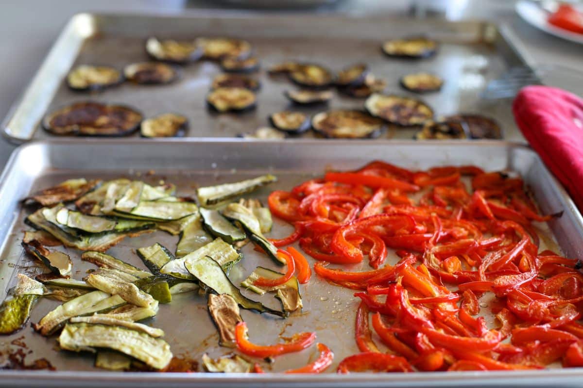 Roasted vegetables on baking sheets: sliced eggplant, zucchini, and red bell peppers arranged in separate sections.