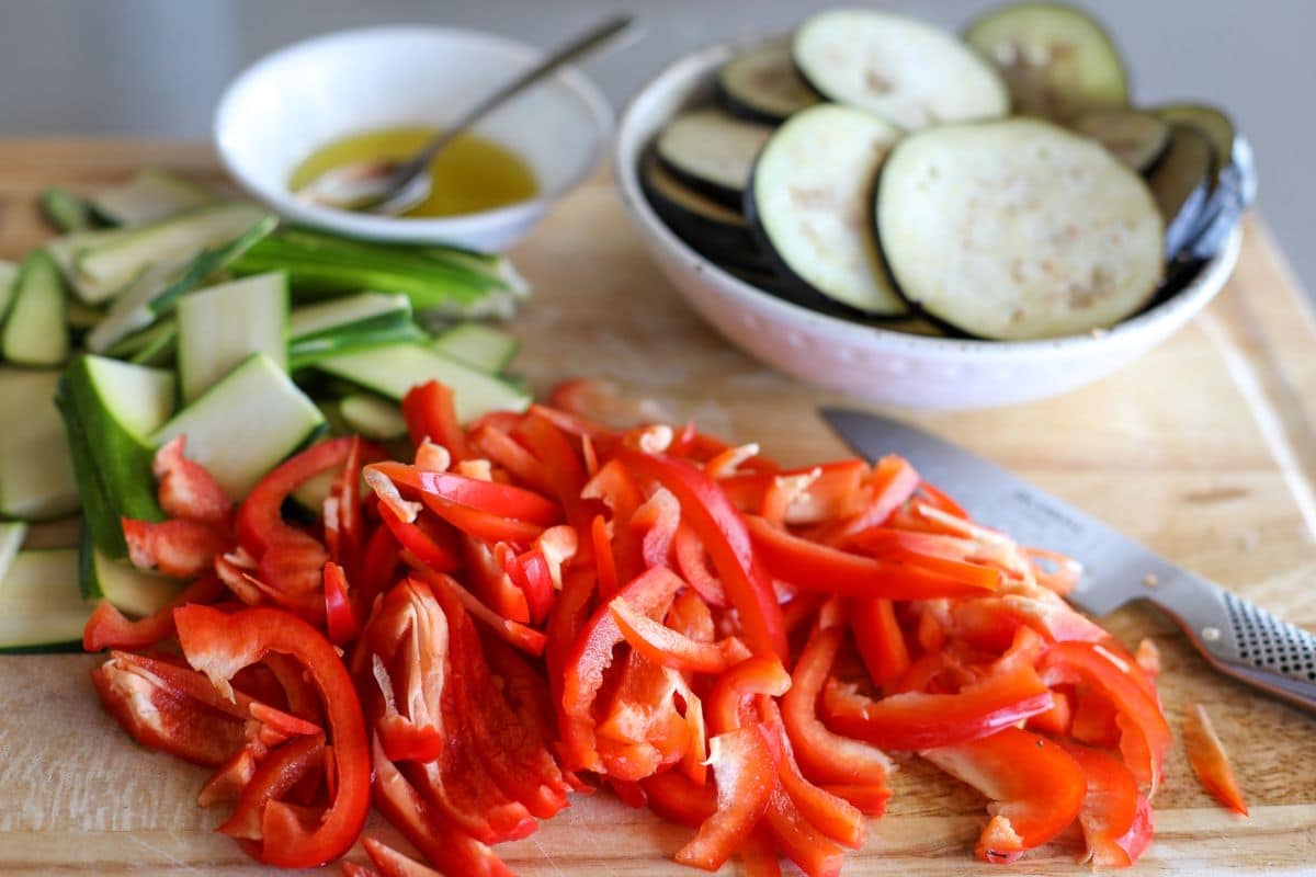 Sliced red bell peppers, zucchini, and eggplant on a cutting board with a knife; bowl of olive oil and eggplant in the background.