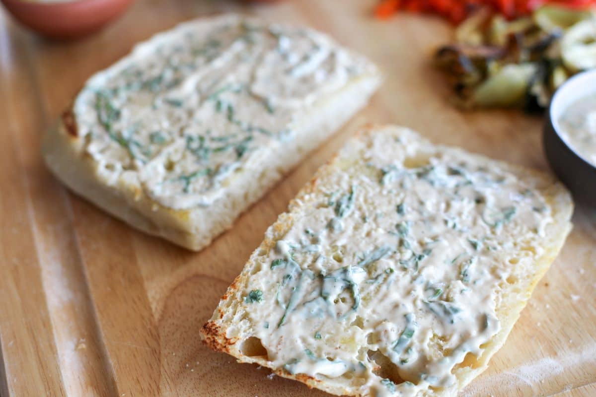 Two slices of bread are spread with a creamy, herb-filled mixture on a wooden surface, with some roasted vegetables visible in the background.