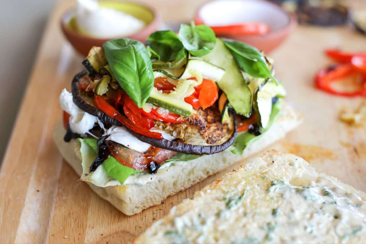 Open-faced sandwich with grilled eggplant, zucchini, roasted peppers, fresh basil, mozzarella, and lettuce on ciabatta bread, with a spread visible on the bread slice.