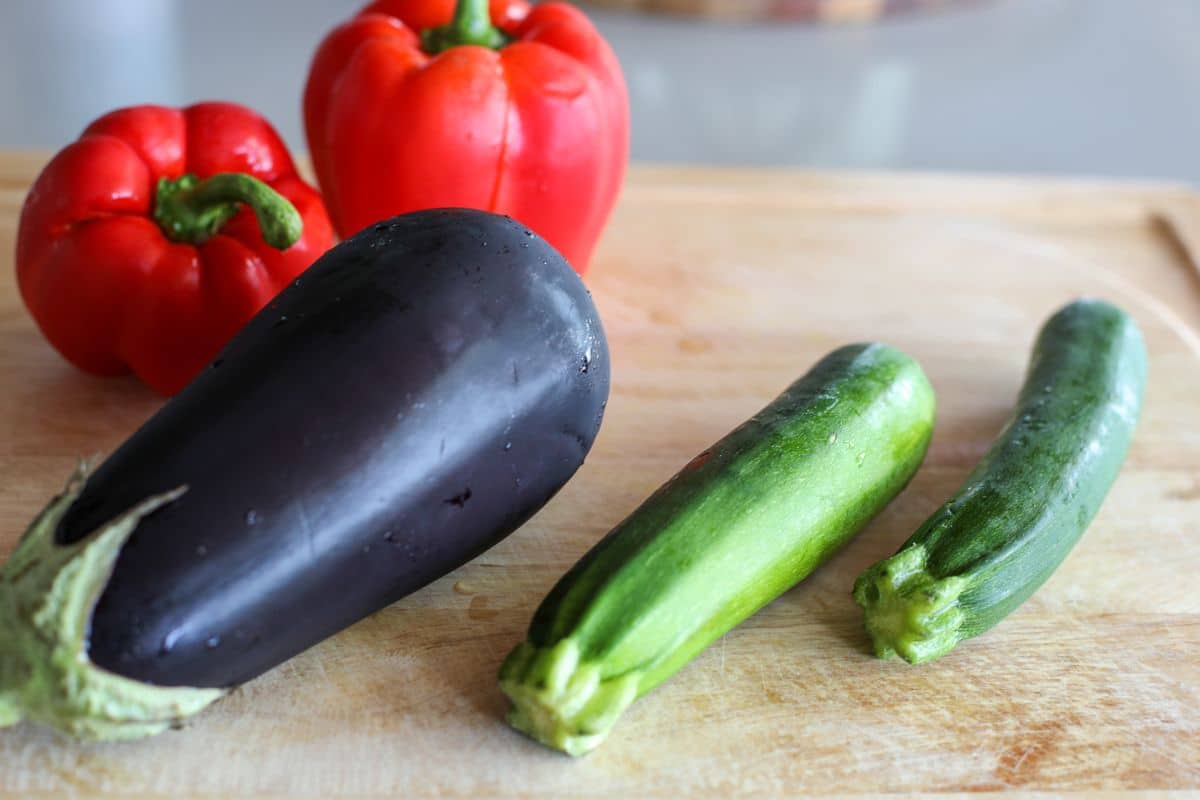 A whole eggplant, two zucchinis, and two red bell peppers on a wooden cutting board.