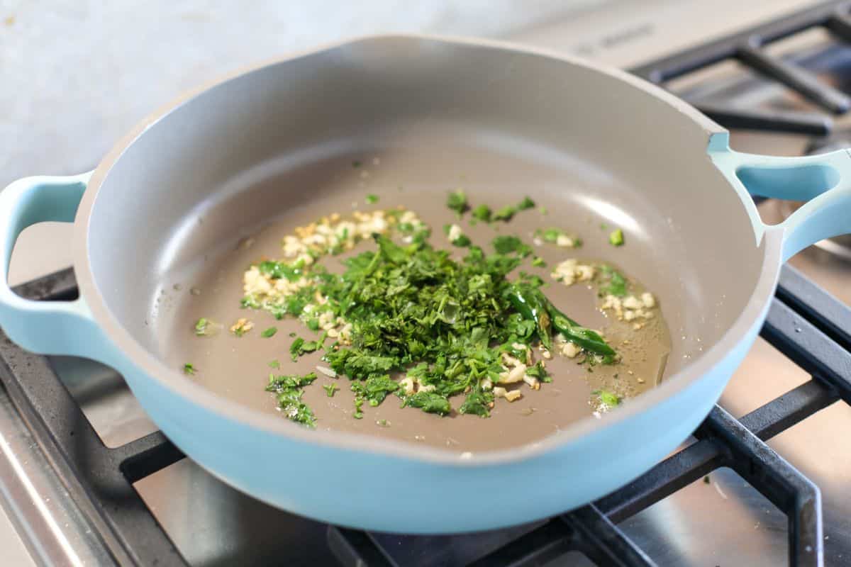 Fresh cilantro and minced garlic sauté in oil in a light blue skillet on a gas stove.