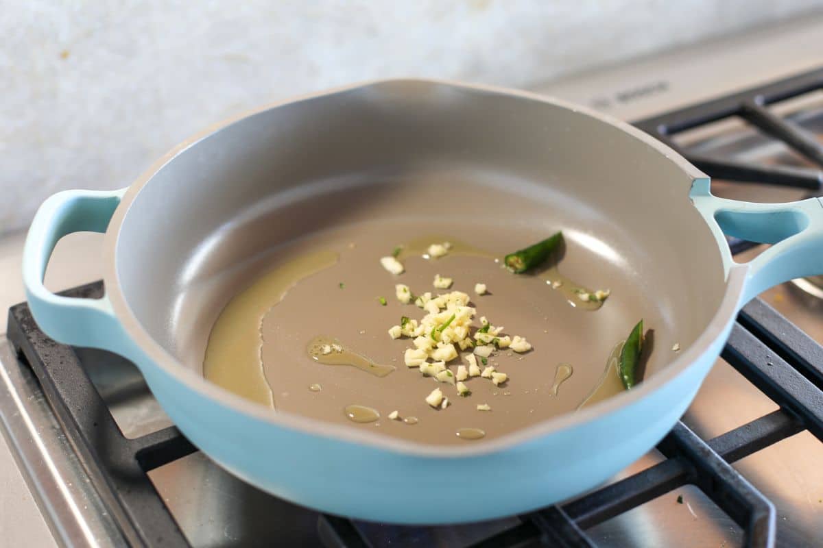 A light blue frying pan on a gas stove contains oil, chopped garlic, and green chili pieces.