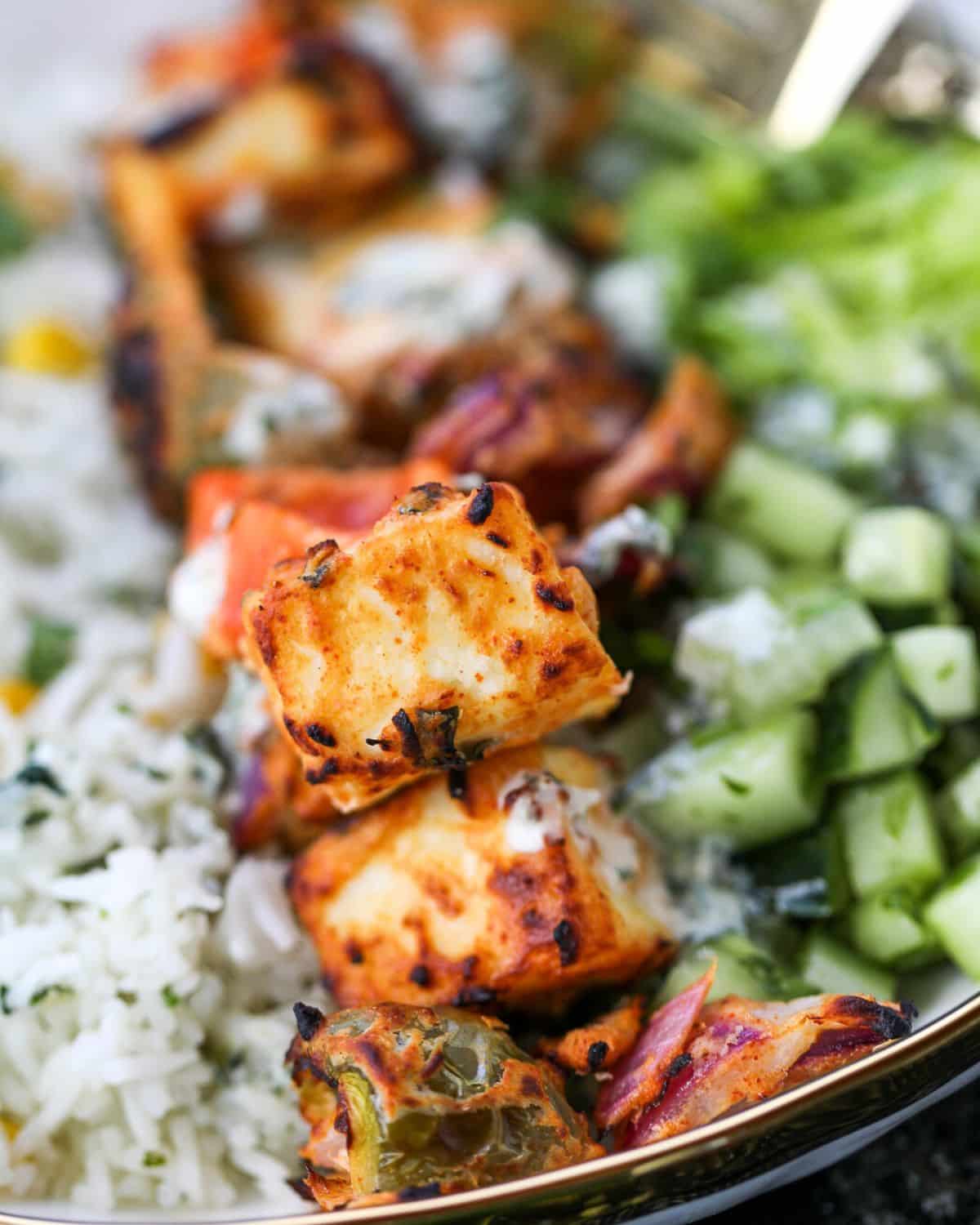 Close-up of grilled paneer tikka and veggies served with white rice, chopped cucumber, and greens on a plate.