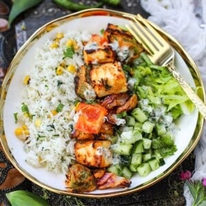 A bowl of Paneer Tikka Rice Bowl with corn, grilled paneer, bell peppers, chopped cucumber, lettuce, and a drizzle of yogurt sauce, served with a gold fork.