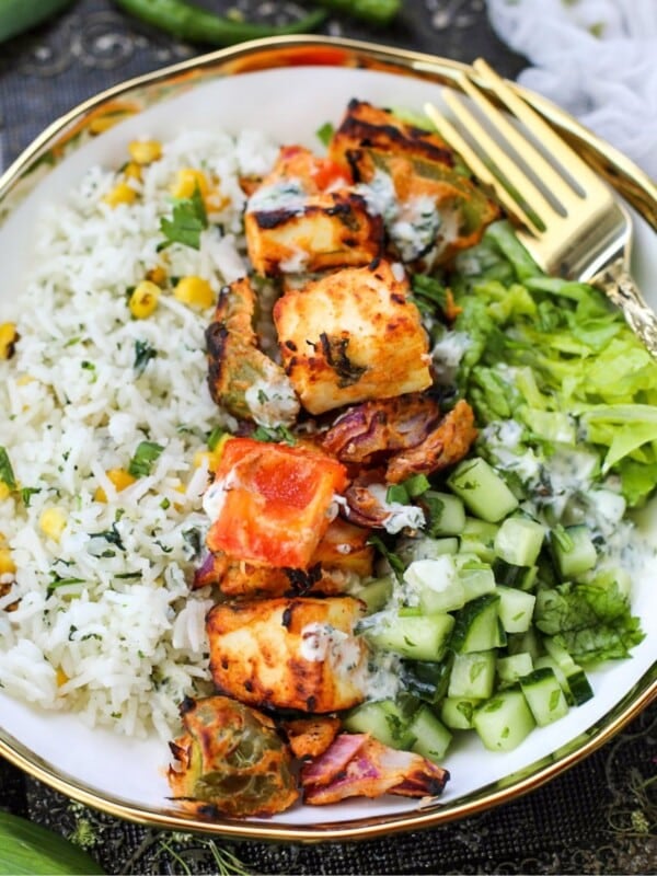 A bowl of Paneer Tikka Rice Bowl with corn, grilled paneer, bell peppers, chopped cucumber, lettuce, and a drizzle of yogurt sauce, served with a gold fork.
