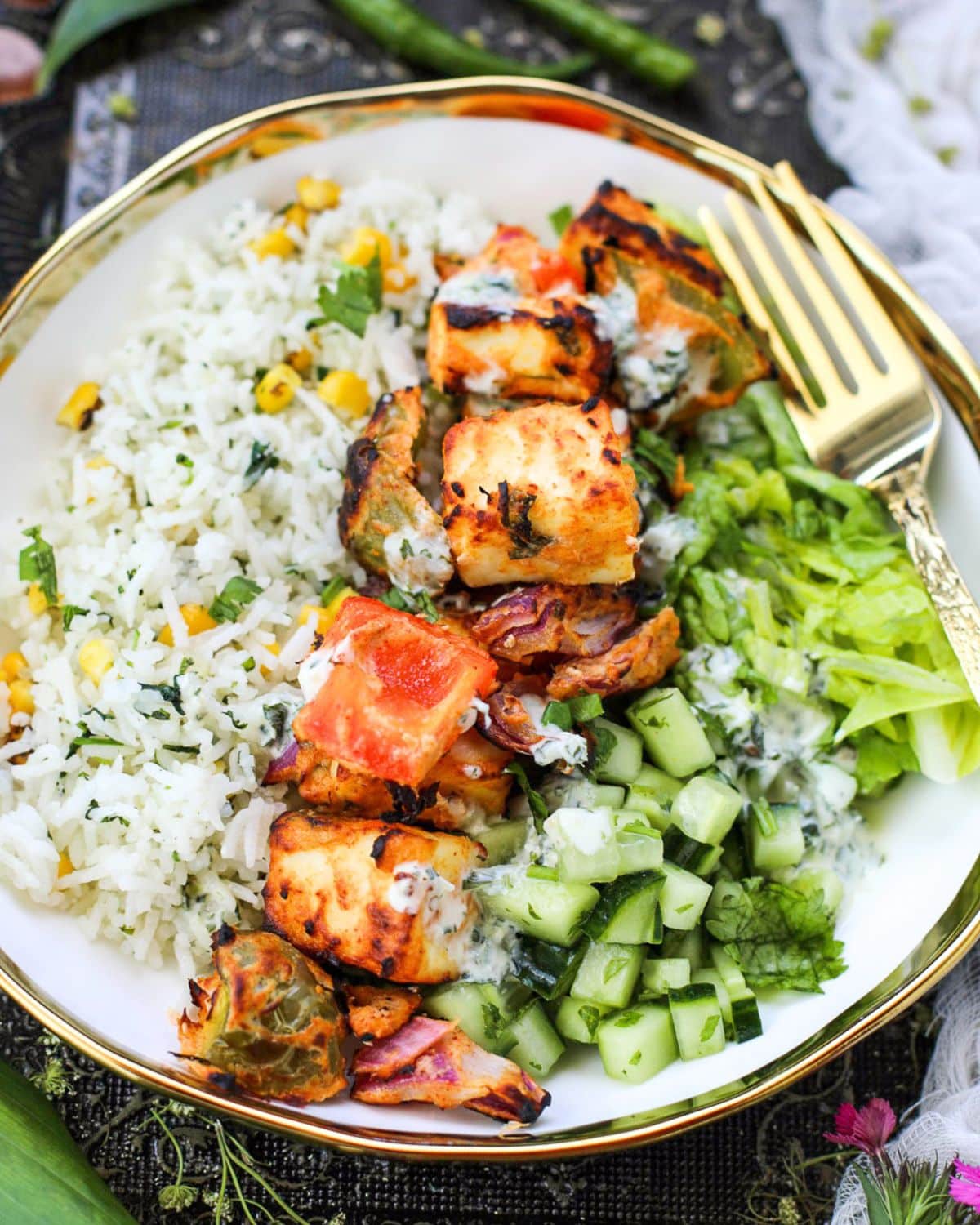 A bowl of Paneer Tikka Rice Bowl with corn, grilled paneer, bell peppers, chopped cucumber, lettuce, and a drizzle of yogurt sauce, served with a gold fork.