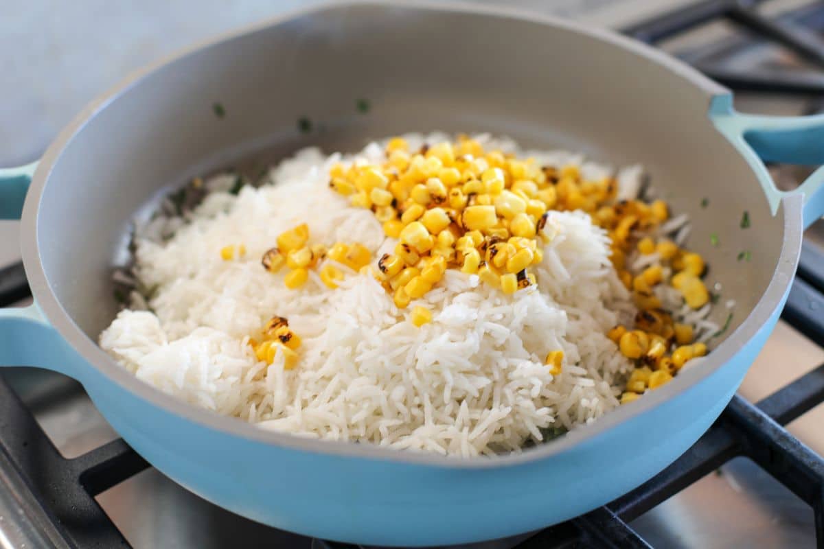 A blue pan on a stove contains cooked white rice topped with fire-roasted corn kernels.