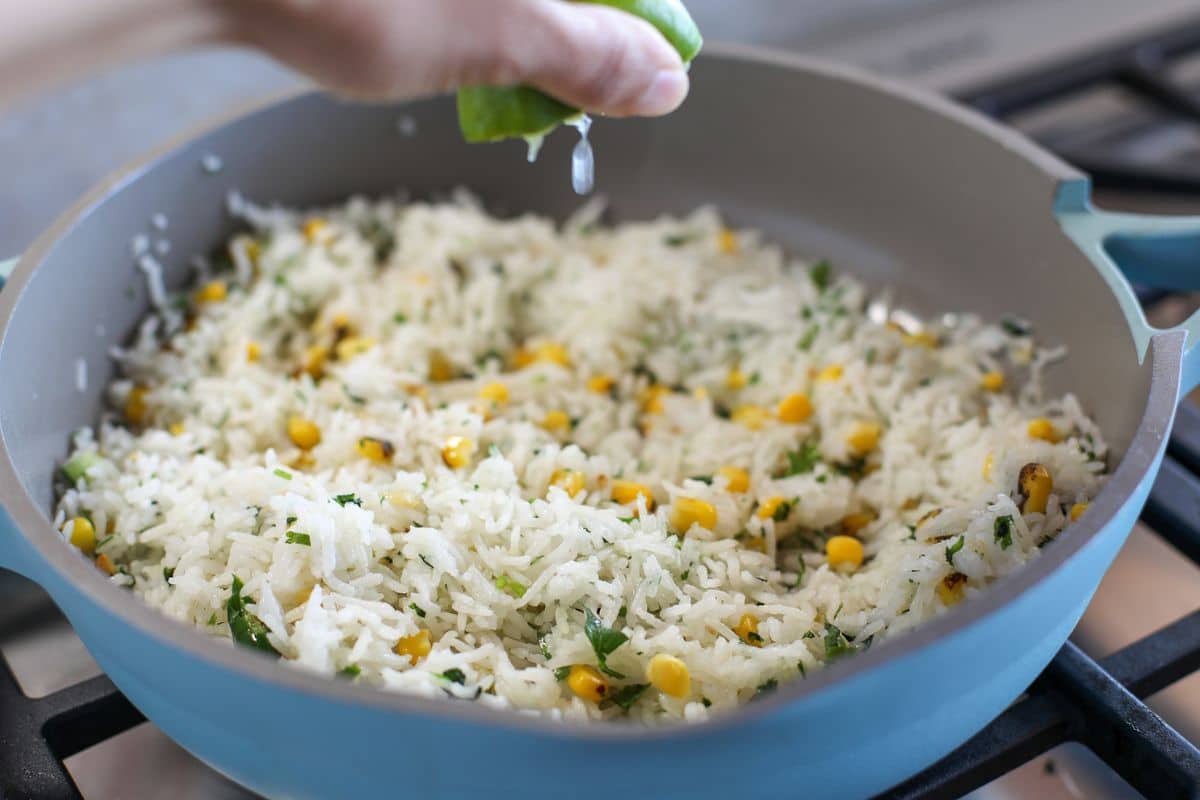 A hand squeezes lime juice over a skillet of cooked rice mixed with corn and chopped herbs on a stovetop.