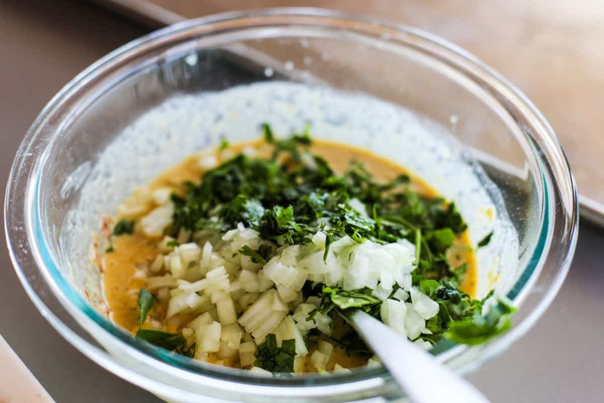 A glass bowl containing a beasn batter topped with chopped onions and spinach.