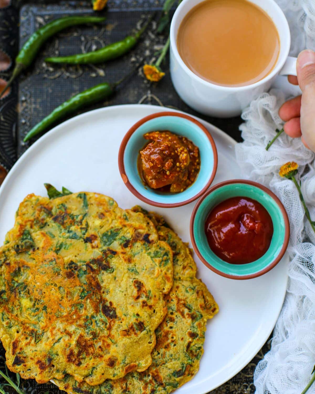 A plate with two savory pancakes, served with bowls of pickle and ketchup, a cup of tea, and green chilies on the side.