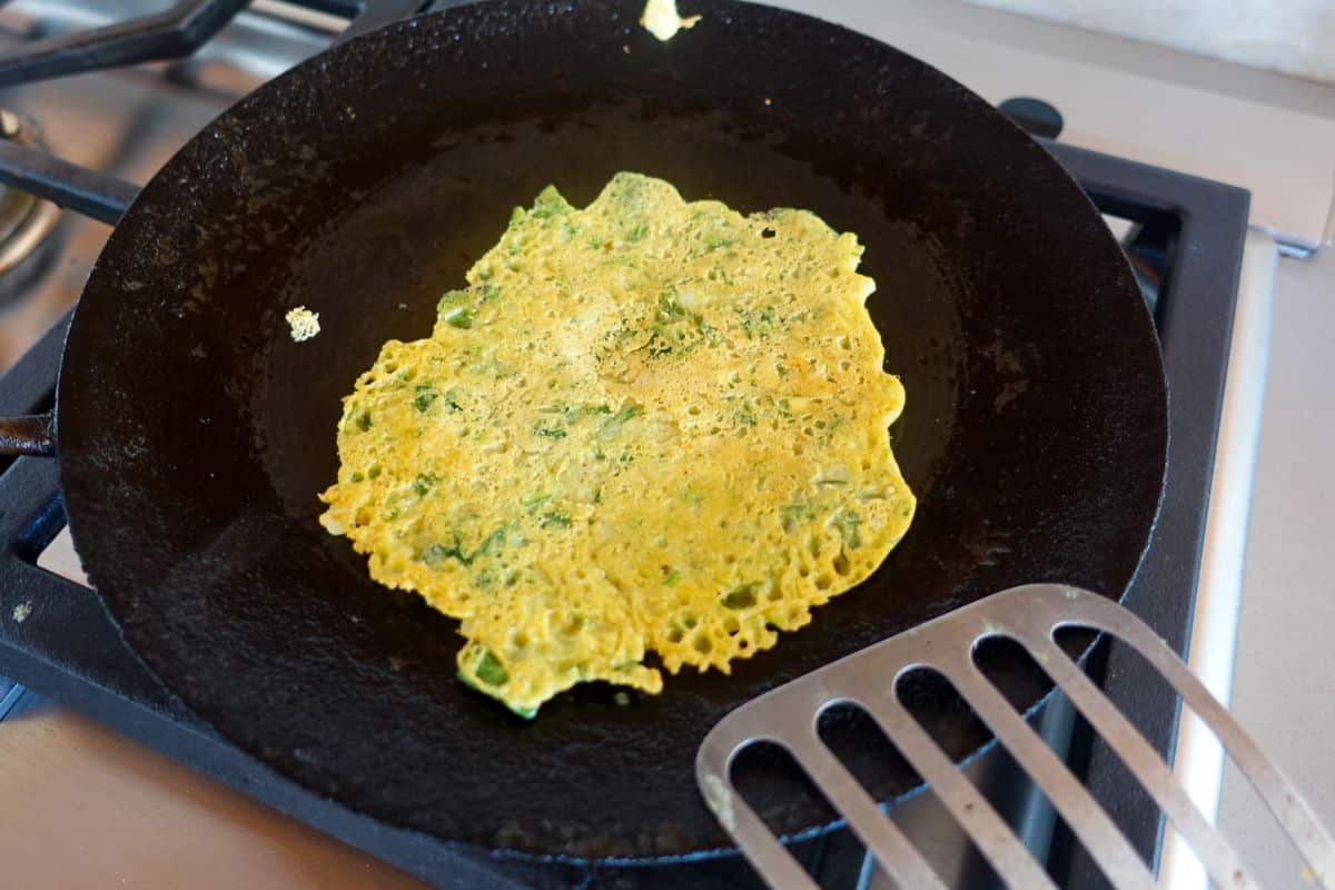 A thin besan chilla with green herbs cooks in a black frying pan on the other side.