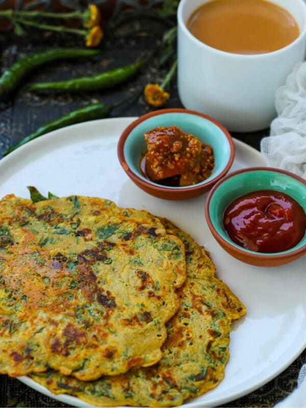 A white plate with two savory pancakes, a small bowl of pickle, and a bowl of red sauce, beside a cup of tea, green chilies, and yellow flowers on a dark surface.
