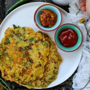 A white plate with two savory pancakes served with small bowls of red sauce and pickle, garnished with a green chili.